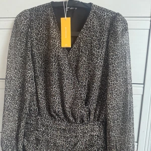 Leopardmönstrad klänning från Gina Tricot - Snygg leopardmönstrad klänning från Gina Tricot med v-ringning och långa ärmar. Perfekt för att sticka ut med ett djärvt mönster. Klänningen har en smickrande passform med rynkdetaljer som ger en fin siluett. 🐆✨