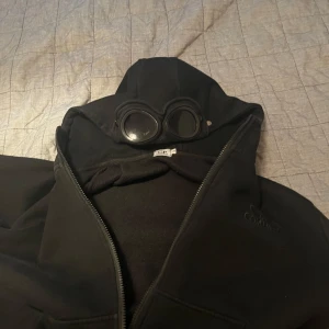 Svart jacka med glasögon från C.P. Company - Säljer en svart jacka från C.P. Company med inbyggda glasögon i huvan. Jackan har dragkedja och är långärmad, perfekt för en unik stil. Märkesloggan är broderad på bröstet.