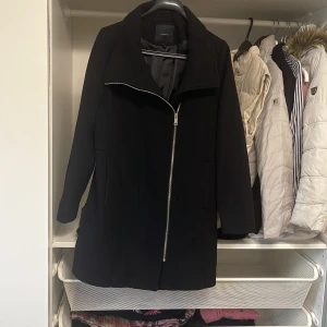 Svart kappa från Esprit - Snygg svart kappa från Esprit med dragkedja framtill. Den har en stilren design och är perfekt för kyligare dagar. Jackan har långa ärmar och en klassisk krage. Passar bra till både vardag och mer uppklädda tillfällen.