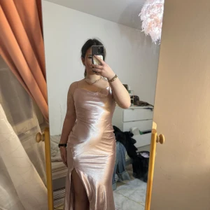 Champagnefärgad långklänning med slits - Elegant champagnefärgad långklänning i satin med smala axelband och en stilren slits framtill. Perfekt för en festlig kväll.. använd 1 gång 