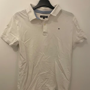 Vit pikétröja från Tommy Hilfiger - Säljer en klassisk vit pikétröja från Tommy Hilfiger med korta ärmar och en liten broderad logotyp på bröstet. Tröjan har en stilren krage med blå detaljer på insidan. Perfekt för en avslappnad och stilren look. Skriv om ni har funderingar och priset är inte hugget i sten!