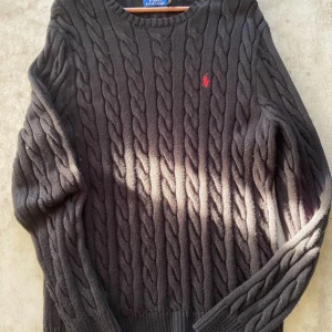 Svart kabelstickad tröja från Ralph Lauren - Säljer en stilren svart kabelstickad tröja från Ralph Lauren med det klassiska röda logotypen på bröstet. Tröjan har långa ärmar och ribbstickade muddar vid ärmslut och nederkant. Perfekt för kyligare dagar!