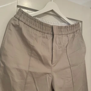 Ljusgrå byxor från Tiger of Sweden - Snygga beige byxor från Tiger of Sweden med elastisk midja för extra komfort. Tillverkade i en blandning av bomull och elastan, skulle beskriva det som en blandning av chinos, kostymbyxa och trackpants