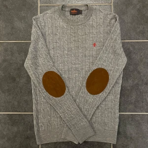 Grå stickad tröja från Morris - Säljer en stilren grå stickad tröja från Morris med kabelstickat mönster och bruna armbågslappar. Tröjan har långa ärmar och rund halsringning, perfekt för kyligare dagar. (100% merinoull)