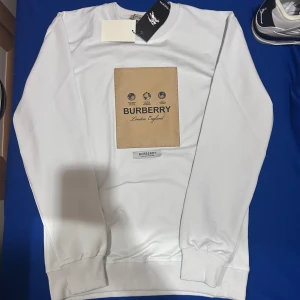 Vit långärmad tröja från Burberry - Säljer en stilren vit långärmad tröja från Burberry med ett stort beige tryck på bröstet. Tröjan har ribbade muddar vid ärmslut och nederkant. Perfekt för en avslappnad och trendig look.