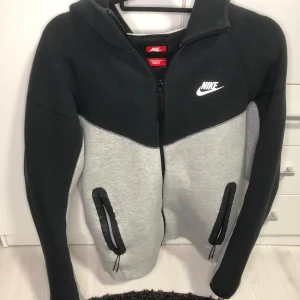 Svart och grå nike tech zip hoodie - Säljer en snygg svart och grå hoodie från Nike med dragkedja och huva. Jackan har två praktiska fickor med dragkedjor och en klassisk Nike-logga på bröstet. Perfekt för en sportig look eller en avslappnad dag. Köpt för 1500kr från Nike.