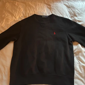 Svart tröja från Ralph Lauren - Säljer en klassisk svart tröja från Ralph Lauren med det ikoniska röda logotypbroderiet på bröstet. Tröjan har långa ärmar och en rund halsringning. Perfekt för en stilren look. 