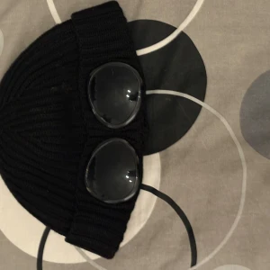 C.P company mössa  - Snygg svart ribbad mössa från C.P. Company med unika glasögondetaljer framtill. Perfekt för att ge din outfit en cool touch. Tillverkad i ett mjukt och bekvämt material.