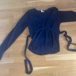 Mörkblå omlottopp från Nelly - Säljer en elegant mörkblå omlottopp från Nelly. Toppen har långa ärmar och ett knytband i midjan för en smickrande passform. Perfekt för både vardag och speciella tillfällen.