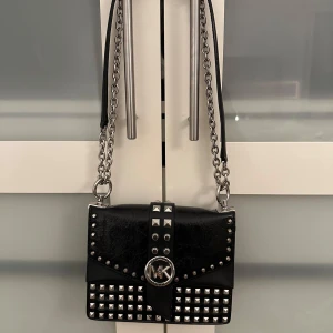 Svart från Michael Kors - Snygg svart väska från Michael Kors med coola nitar. Väskan har ett stort MK-emblem. Tillverkad i skinn med detaljer som ger en lyxig känsla. Originalpris: 4000kr