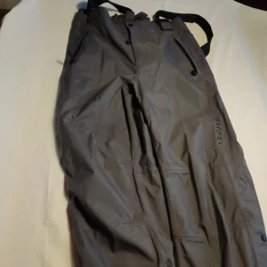 Grå täckbyxor från Craft - Säljer ett par grå täckbyxor från Craft i storlek L. Byxorna har justerbara hängslen och dragkedjor vid fickorna. Perfekta för kalla dagar med sin robusta design.