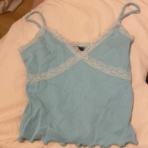 Ljusblå ribbad linnetopp från Gina Tricot - Säljer en söt ljusblå ribbad linnetopp från Gina Tricot. Toppen har spetsdetaljer vid halsringningen och tunna axelband. Perfekt för varma dagar eller som ett lager under en tröja.