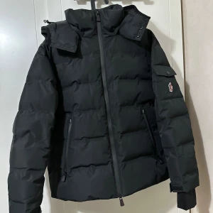 Svart dunjacka från Moncler - Säljer en stilren svart dunjacka från Moncler med dragkedja och broderad logga på ärmen. Jackan har en hög krage och avtagbar huva, perfekt för kyliga dagar. Den är designad för att hålla dig varm och bekväm med sin quiltade stil.
