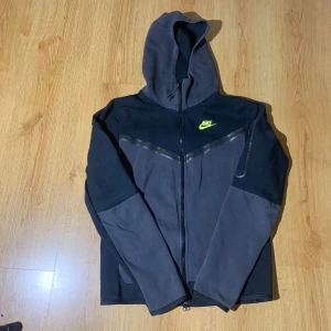 Nike tech  - Säljer en svart hoodie från Nike med dragkedja och huva. Den har en gul Nike-logga på bröstet och är tillverkad i ett mjukt material. Perfekt för en sportig look eller avslappnade dagar.