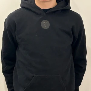 Svart hoodie från Trace - Säljer en svart hoodie från Trace med en stor logga på ryggen och en mindre logga på framsidan. Perfekt för en avslappnad stil. Hoodien har en klassisk passform med långa ärmar och en bekväm huva. Skriv om ni har funderingar och priset är inte hugget i sten!