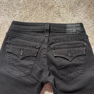 Svarta true religon jeans med nitar - Sjukt snygga svarta, Lågmidjade bootcut jeans från True Religion med coola nitdetaljer på fickorna. Innerbenslängden är 85 cm och midjemåttet tvärsöver är 35-36 cm. Jeansen passar någon med storlek XXS-XS. Jag på bilden är 168 cm