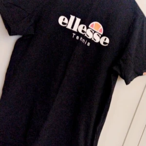 Svart t-shirt från Ellesse - Snygg svart t-shirt från Ellesse med deras ikoniska logga och texten 'Tennis' på framsidan. Perfekt för en sportig look eller avslappnad stil. Kortärmad och bekväm passform. Strl L