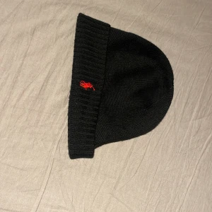 Svart mössa från Polo Ralph Lauren - Snygg svart mössa från Polo Ralph Lauren i merinoull. Den har en röd broderad logga framtill och är perfekt för kyliga dagar. Mössan är ribbad och ger en stilren look. Qr koden kan skannas