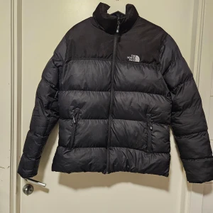 Svart dunjacka  - Säljer en svart dunjacka med dragkedja och två fickor framtill. Jackan har en hög krage och är perfekt för kalla dagar. Den är märkt med '700' på ärmen, vilket indikerar dess fyllningsgrad.