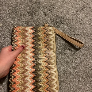 Mönstrad kuvertväska med dragkedja - Snygg kuvertväska med ett färgglatt zigzag-mönster i orange, beige och brunt. Väskan har en praktisk dragkedja och en handledsrem för enkel bärning. Perfekt för att ge en färgklick till din outfit!