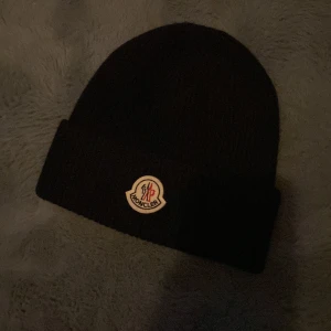 Svart mössa från Moncler - Snygg svart mössa från Moncler med broderad logotyp framtill. Den är ribbad och stickad, perfekt för kyliga dagar. En stilren accessoar som håller dig varm och trendig.