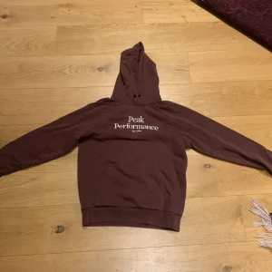 Vinröd hoodie från peak performance  - Säljer en vinröd hoodie från Peak Performance med vit logga på bröstet. Tröjan har en klassisk design med huva och en stor ficka framtill. Skick 9/10, Inga slitningar eller tecken på användning 