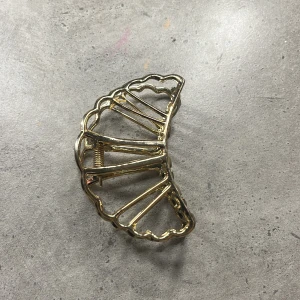 Guldfärgad hårklämma i metall - Elegant guldfärgad hårklämma i metall med ett vackert, böljande mönster. Perfekt för att hålla håret på plats med stil. Klämman har en robust fjädermekanism för säker passform.