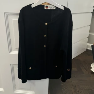 Svart kofta från Busnel - Elegant svart kofta från Busnel med guldknappar. Koftan har två designfickor fram och guldknappar både på armarna och i knäppningen fram. Storlek framgår inte men passar S/M. Bra skick!