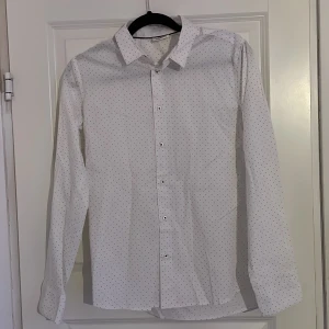 Vit prickig skjorta - Snygg vit skjorta med små prickar och klassisk krage. Perfekt för en stilren look. Skjortan har långa ärmar och knappar framtill. Storlek 170