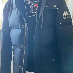 Marinblå dunjacka från Moose Knuckles - Extravagant marinblå dunjacka från Moose Knuckles med dragkedja och knappar. Jackan har en stilren design med en logotyp på ärmen och är perfekt för kyliga dagar. Den är utrustad med NFC-teknologi för extra funktionalitet.