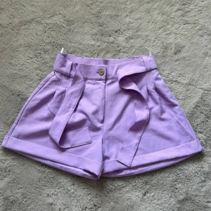 Lila shorts med knytband - Snygga lila shorts med hög midja och knytband. Perfekta för sommardagar, med en avslappnad passform och uppvikta ben. De har en knapp framtill och praktiska fickor.