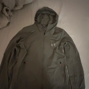 Grå vindjacka från Under Armour - Säljer en grå vindjacka från Under Armour med huva och dragkedja. Jackan är perfekt för blåsiga dagar. Den har en stilren design med logotyp på bröstet. Jackan är i storlek M men passar garanterat som bäst på dig som har storlek xs-s i tunnare jackor. (Priset kan diskuteras)