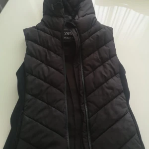 Svart dunväst från Zara - Snygg svart dunväst från Zara med dragkedja. Perfekt för kyligare dagar med sin quiltade design som ger extra värme och stil. Passar bra till både casual och mer uppklädda tillfällen.