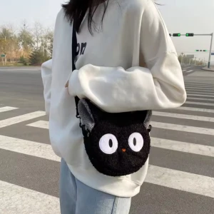Svart mini-axelväska med kattmotiv, Jiji från Kickis expressbud, Ghibli - Söt och unik liten svart axelväska i form av en katt med stora vita ögon och gråa öron. Katten är Jiji från Kickis expressbud från Ghibli Studios. Perfekt för att ge en lekfull touch till din outfit. Väskan har en justerbar axelrem och är tillverkad i ett mjukt material.