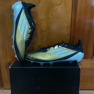 Adidas f50 - Snygga fotbollsskor från Adidas i guld och svart med vita detaljer. Skorna har snörning och är designade för optimal prestanda på planen. Perfekta för fotbollsentusiaster som vill sticka ut.