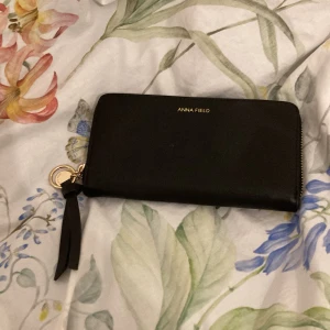 Svart plånbok från Anna Field - Snygg och stilren svart plånbok från Anna Field med dragkedja och en praktisk rem. Perfekt för att hålla ordning på kort och kontanter. Enkel och elegant design som passar till alla tillfällen. Köpt för 169 och är använd ett par gånger