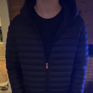 Svart pufferjacka från Save the Duck - Säljer en svart pufferjacka från Save the Duck i storlek M. Jackan är 100% djurfri och har en dragkedja framtill. Perfekt för kyliga dagar med sin quiltade design och långa ärmar. Den har en cool orange logga på ärmen. Passar både höst och vinter! 