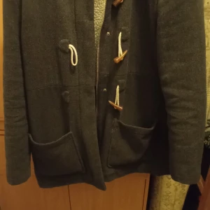 Grå duffeljacka från Red Label - Säljer en snygg grå duffeljacka från Red Label med träknappar och snördetaljer. Jackan har en mysig fleecefoder och praktiska fickor framtill. Mjukt material 