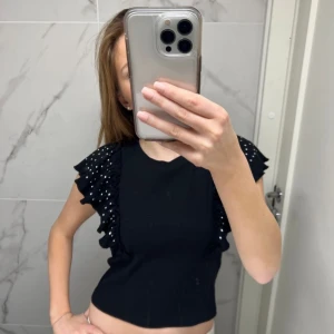 Svart topp med volangdetaljer - Snygg svart topp med volangdetaljer på ärmarna. Perfekt för en stilren look. Toppen är kortärmad och har en tight passform. Storlek 170 men passar 34,36,38💞