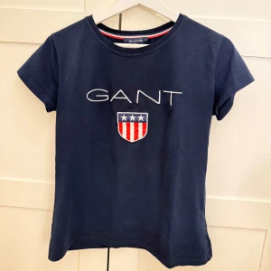 Mörkblå t-shirt från GANT - Snygg mörkblå t-shirt från GANT med ett broderat emblem och text på bröstet. Perfekt för en avslappnad stil med korta ärmar och rund halsringning. Jätte bra skick, är liten i storlek så passar mer som M