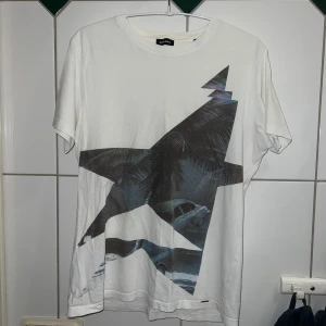 Vit t-shirt med grafiskt tryck från Diesel - Säljer en vit t-shirt från Diesel med ett coolt grafiskt tryck i svart och blått. Perfekt för en avslappnad stil. T-shirten är kortärmad och gjord i mjukt material.