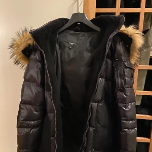 Svart dunjacka med pälsdetaljer - Snygg svart dunjacka med pälskantad huva. Jackan har en glansig finish och flera praktiska fickor med dragkedja. Perfekt för kalla dagar med stil.