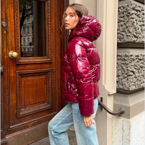 Vinröd pufferjacka med huva - Snygg och glansig vinröd pufferjacka med huva och dragkedja. Perfekt för vintern! Säljer då den inte kommer till användning, den är i nyskick! 