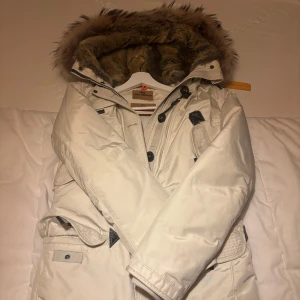 Vit parkajacka med päls - Snygg vit parkajacka från Parajumpers med avtagbar äkta pläs på huvan. Jackan har flera praktiska fickor och stängs med både dragkedja och knappar. Perfekt för kalla vinterdagar.