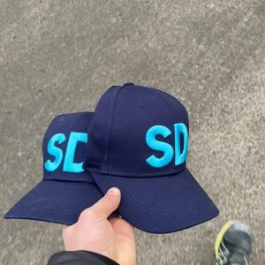 Mörkblå keps med turkos text - Snygg mörkblå keps med stora turkosa bokstäver 'SD' framtill. Perfekt för en casual look eller som en statement-accessoar. Justerbar passform för optimal komfort.100kr st 