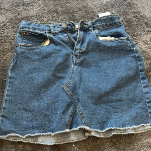 Blå jeanskjol från Levi's - Snygg blå jeanskjol från Levi's med klassisk femficksdesign och rå kant. Kjolen har en knapp och dragkedja framtill samt märkets ikoniska läderpatch baktill. Perfekt för en avslappnad stil.