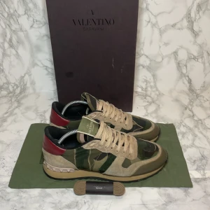 Valentino rockrunner storlek 40 - Snygga Valentino Rockrunner sneakers med ett coolt camouflagemönster i grönt och beige. De är i storlek 39,5 men passar 40 Skorna har röda detaljer och klassiska nitar på hälen. Perfekta för en trendig look. Box ingår ej