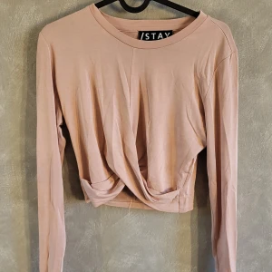 Rosa croppad topp från STAY - Säljer en stilren rosa croppad topp från STAY med långärmad design. Toppen har en unik twistdetalj framtill som ger en modern look. Perfekt för att uppdatera garderoben med något trendigt och bekvämt.