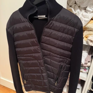 Svart moncler cardigan   - Snygg svart cardigan från Moncler med dragkedja och ribbade detaljer. Jackan har långa ärmar och två praktiska fickor med dragkedja framtill. Perfekt för kyligare dagar. Nfc Scan funkar, använd en handfull gånger max, storlek L sitter men sitter som m