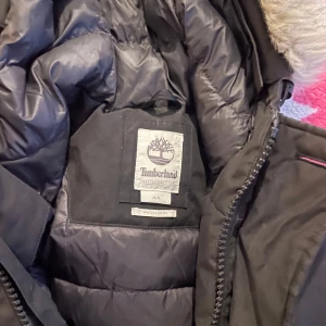 Svart dunjacka från Timberland - Säljer en svart dunjacka från Timberland med pälskantad huva. Jackan har dragkedja och knappar framtill samt flera praktiska fickor. Perfekt för kalla dagar med sin varma och vattentäta design. Har en liten slitage på insidan. 